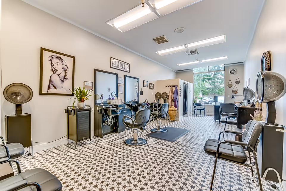Interior of a hair salon with multiple styling chairs, mirrors, and hair drying stations. The floor has a patterned tile design, and there is a large window at the far end letting in natural light. The walls are decorated with framed pictures and a sign that reads 'GLAMOUR'.
