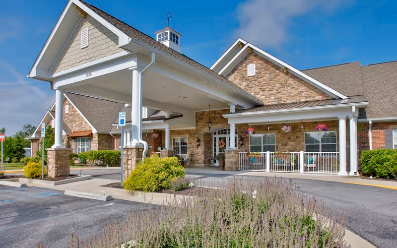 Exterior view of Bickford of Greenwood facility showing a covered entrance with white pillars, stone and brick facade, hanging flower baskets, and a parking area with handicap parking signs.