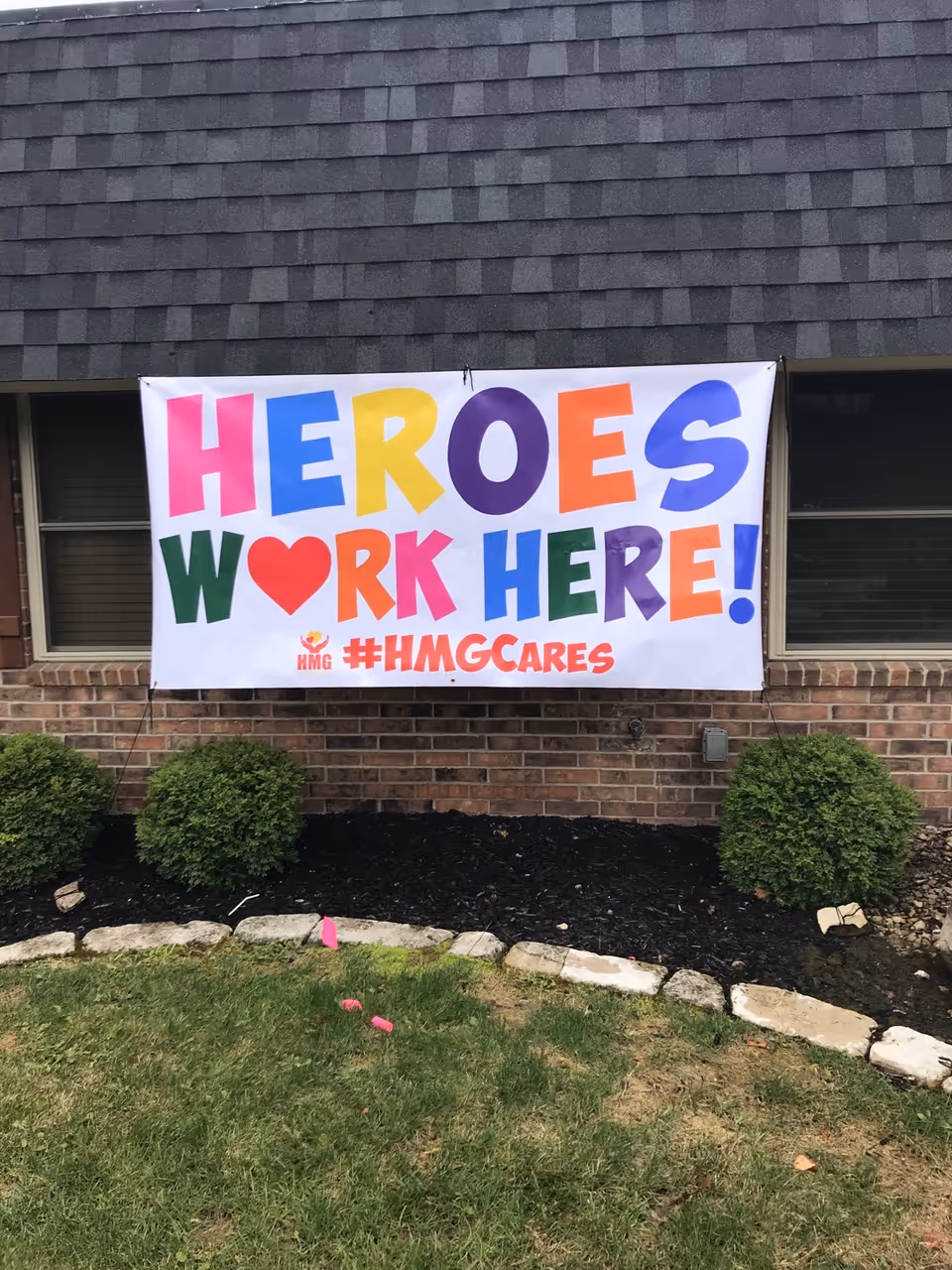 A colorful banner that reads 'HEROES WORK HERE! #HMGCARES' hung on the brick front exterior of a building above shrubs and a small lawn.