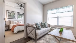 A bright living room with a beige sofa adorned with patterned pillows, a wooden coffee table with green decorative plants, and a large window with a valance letting in natural light. Through an open doorway, a bedroom is visible with a bed featuring white and orange bedding and framed artwork above the headboard.