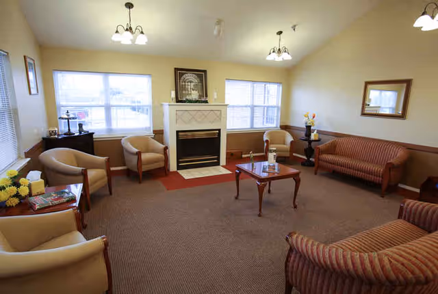 A cozy living room with beige walls and carpet, featuring a fireplace centered between two windows. The room is furnished with several armchairs and sofas arranged around a wooden coffee table. There are decorative items on the mantel and side tables, and ceiling lights provide illumination.