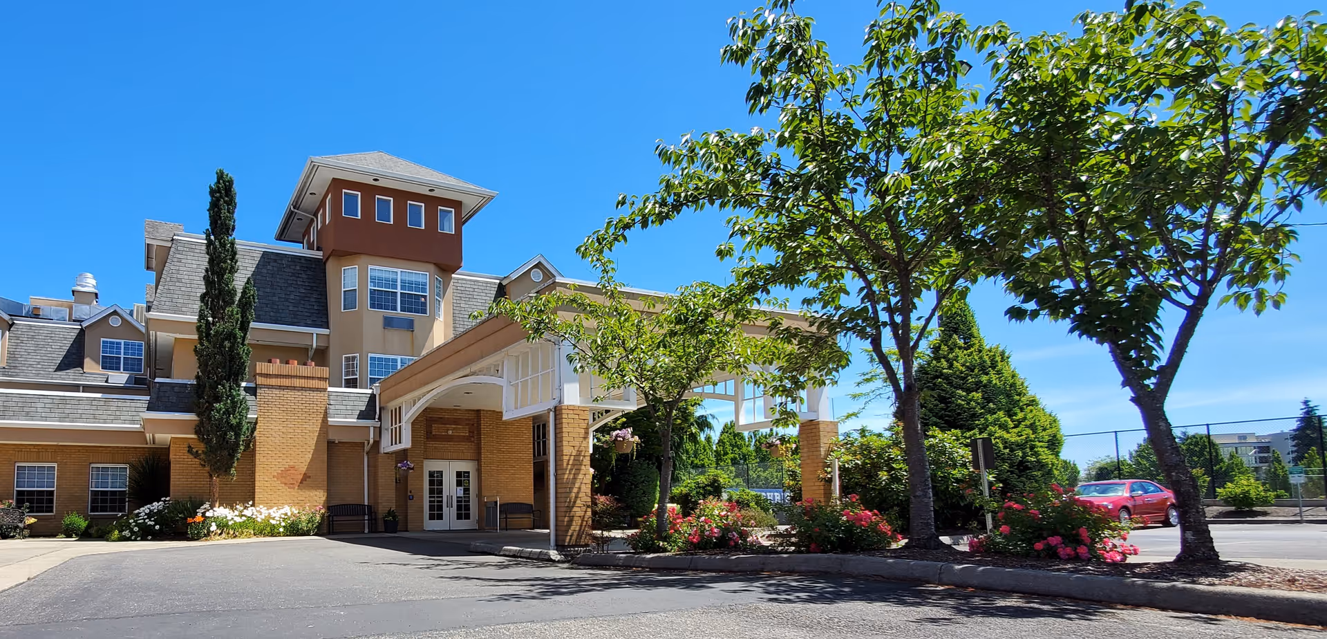 Exterior view of Life Manor Independent Living (Mountainside) building entrance with a covered drop-off area, surrounded by trees, flowering bushes, and a clear blue sky.