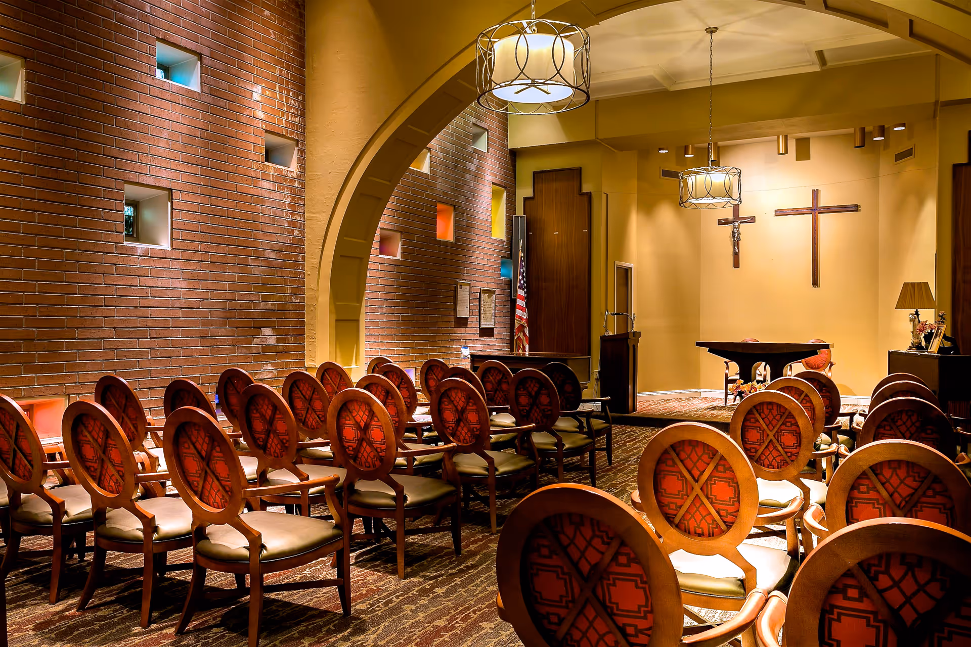 Interior view of a chapel or small worship room with rows of wooden chairs featuring red patterned upholstery, a brick wall with small square windows, two hanging light fixtures, and two crosses mounted on a beige wall behind a small altar table.