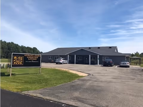 Single-story assisted living building with a parking lot and a lawn sign reading 'Albany Oaks Assisted Living' under a blue sky.