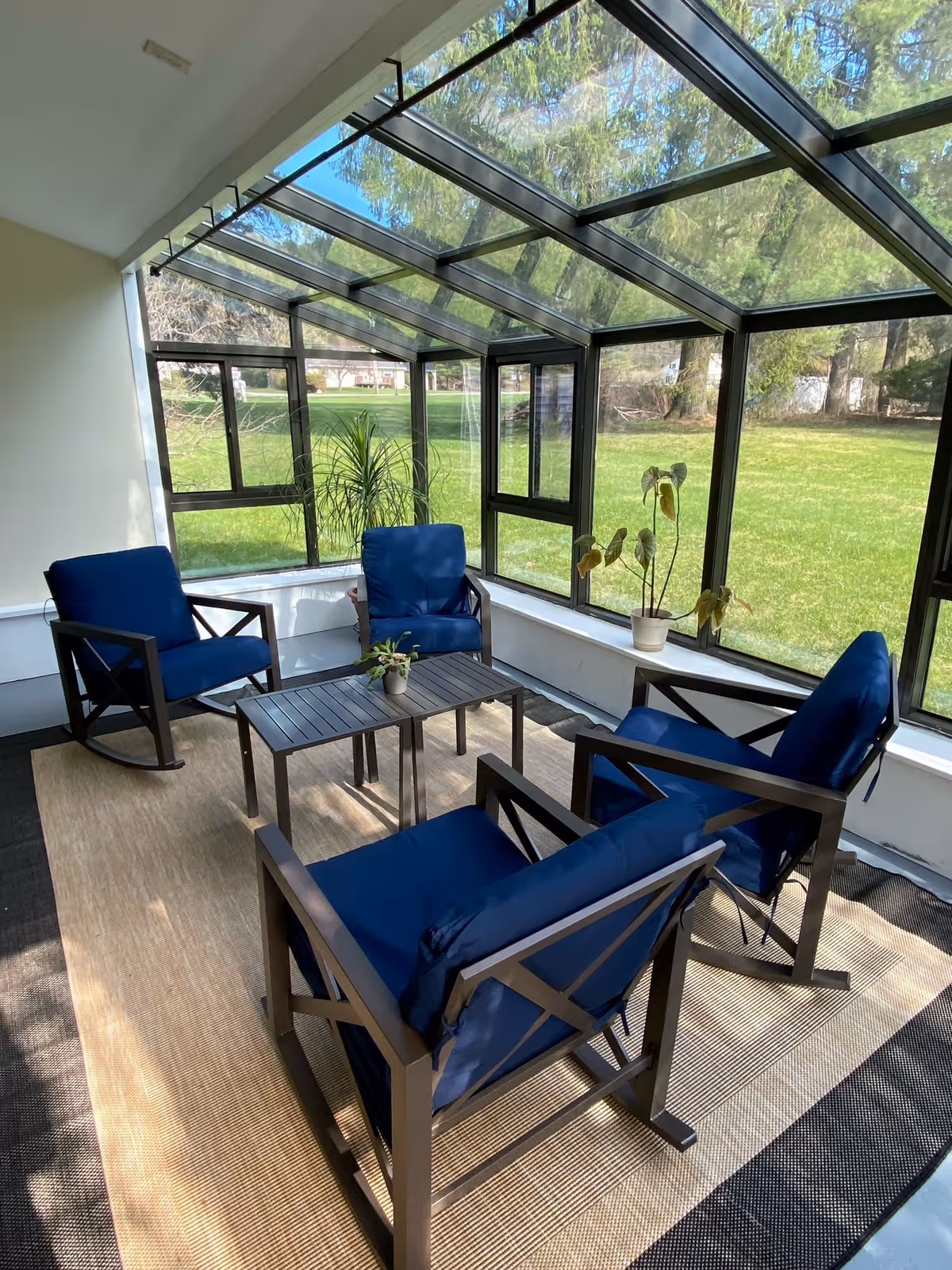 A sunroom with large glass windows and a glass ceiling overlooking a green lawn. The room contains four dark metal-framed rocking chairs with blue cushions arranged around a rectangular black coffee table. There are two potted plants on the window ledge.