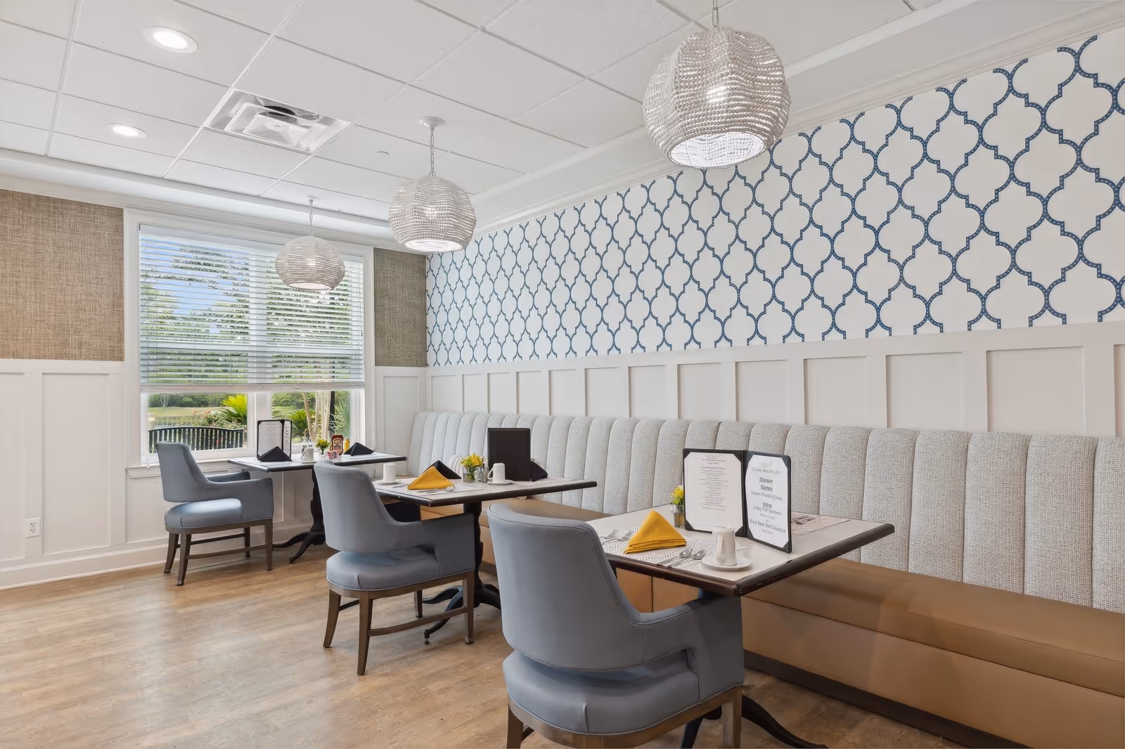 Bright dining area with upholstered banquette seating, tables set with menus and yellow napkins, blue patterned wallpaper, and pendant lights.