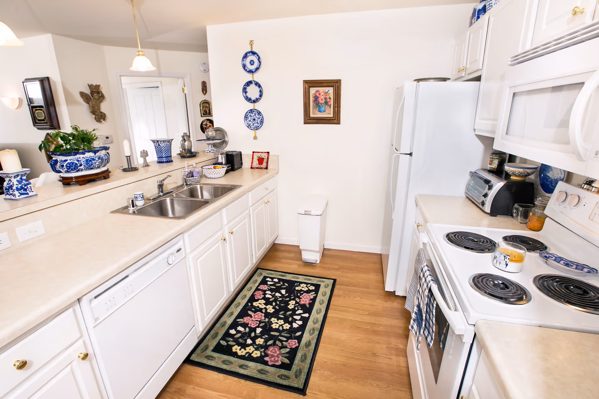 A bright kitchen with white cabinets and appliances including a refrigerator, microwave, stove, and dishwasher. The countertops are beige, and there is a double sink. Decorative blue and white plates hang on the wall, along with a framed floral painting. A small floral rug is placed on the wooden floor, and various kitchen items and plants are arranged on the counter.