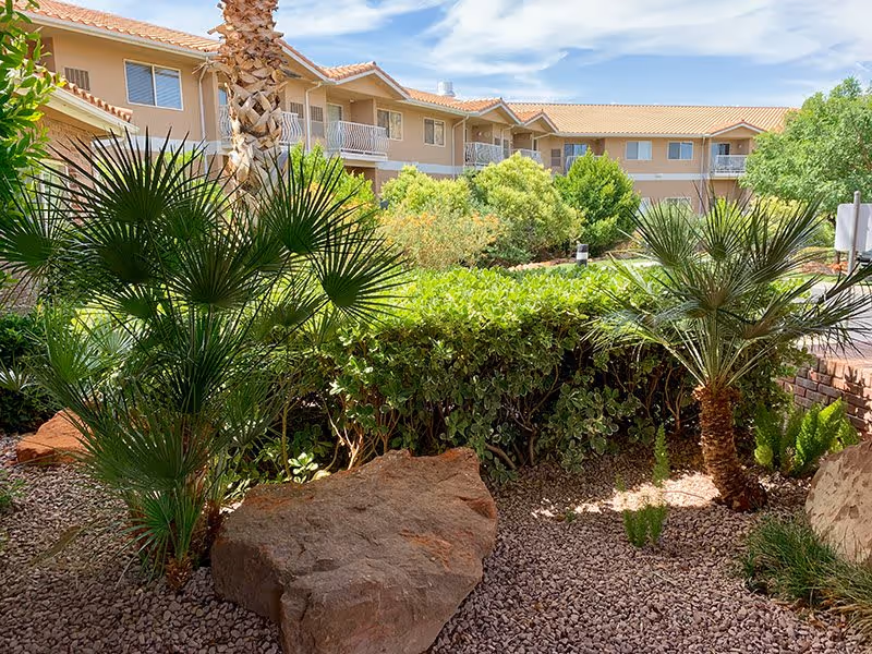 Outdoor garden area with various green plants, palm trees, and large rocks in front of a two-story beige building with balconies under a partly cloudy sky.