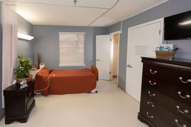A simple and clean bedroom in a healthcare facility with a single bed covered in a brown blanket, a window with closed blinds, a dark wooden dresser with a flat-screen TV mounted above it, and a nightstand with a plant and decorative items. The walls are painted gray and the floor is light-colored.