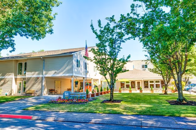 Exterior view of Junction City Retirement and Assisted Living facility showing a two-story building with beige siding, a covered entrance, an American flag on a flagpole, well-maintained green lawn, trees, and flower beds under a clear blue sky.