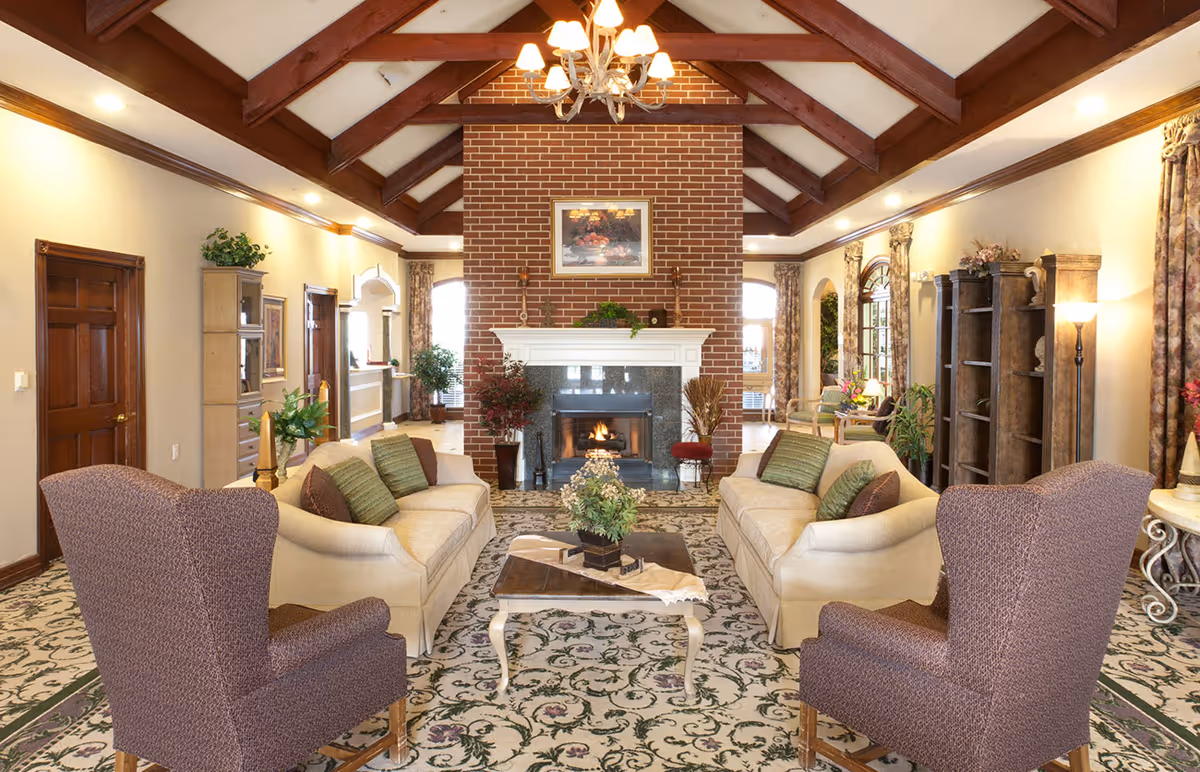 A spacious living room with two beige sofas and two purple armchairs arranged around a coffee table on a patterned carpet. The room features a large brick fireplace with a white mantle, a chandelier hanging from a wooden beam ceiling, and large windows with floral curtains letting in natural light. There are plants and decorative items placed around the room.