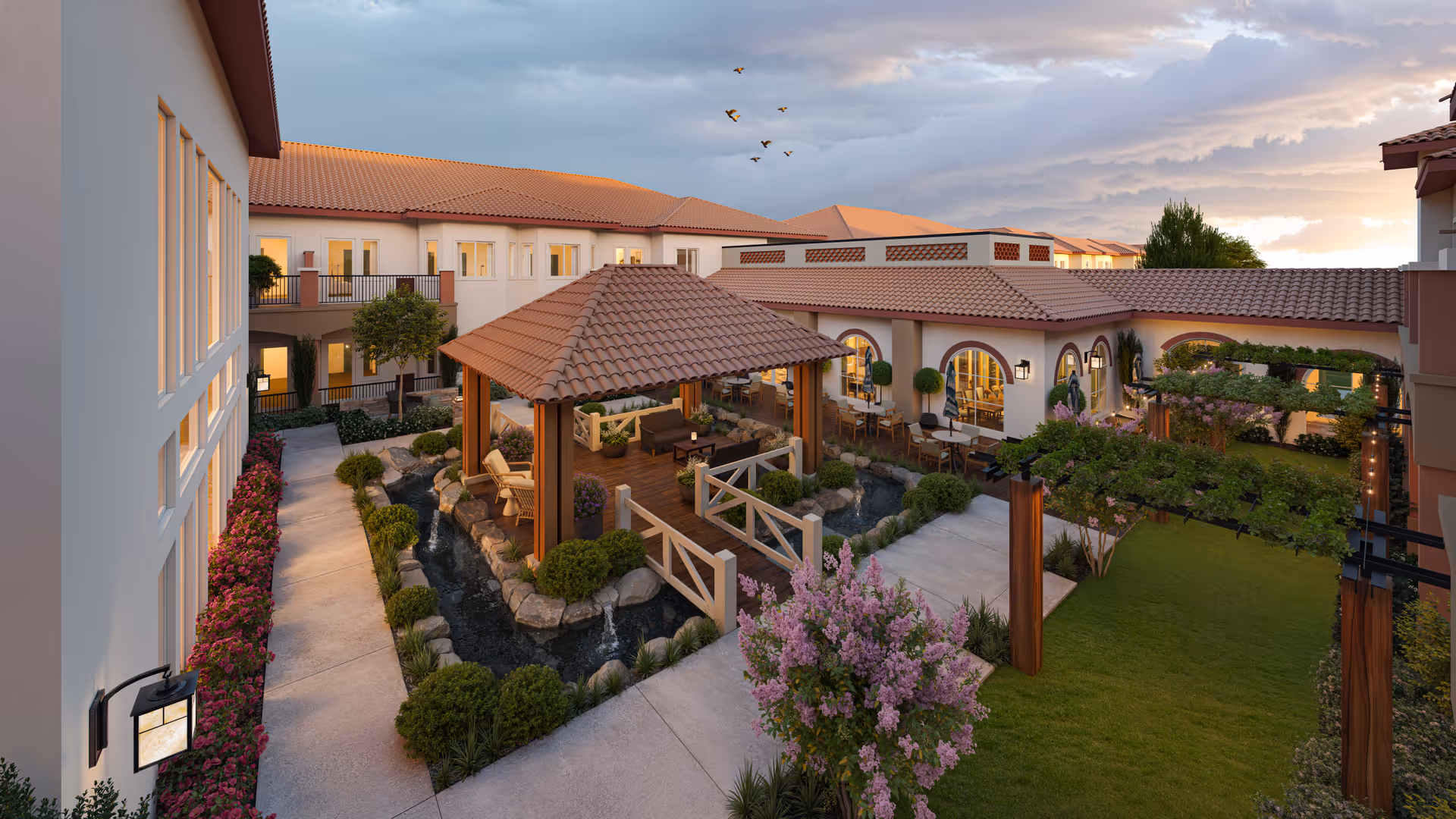 A landscaped central courtyard with a covered gazebo, wooden bridges over a pond, seating and dining areas, and surrounding Mediterranean-style buildings at sunset.