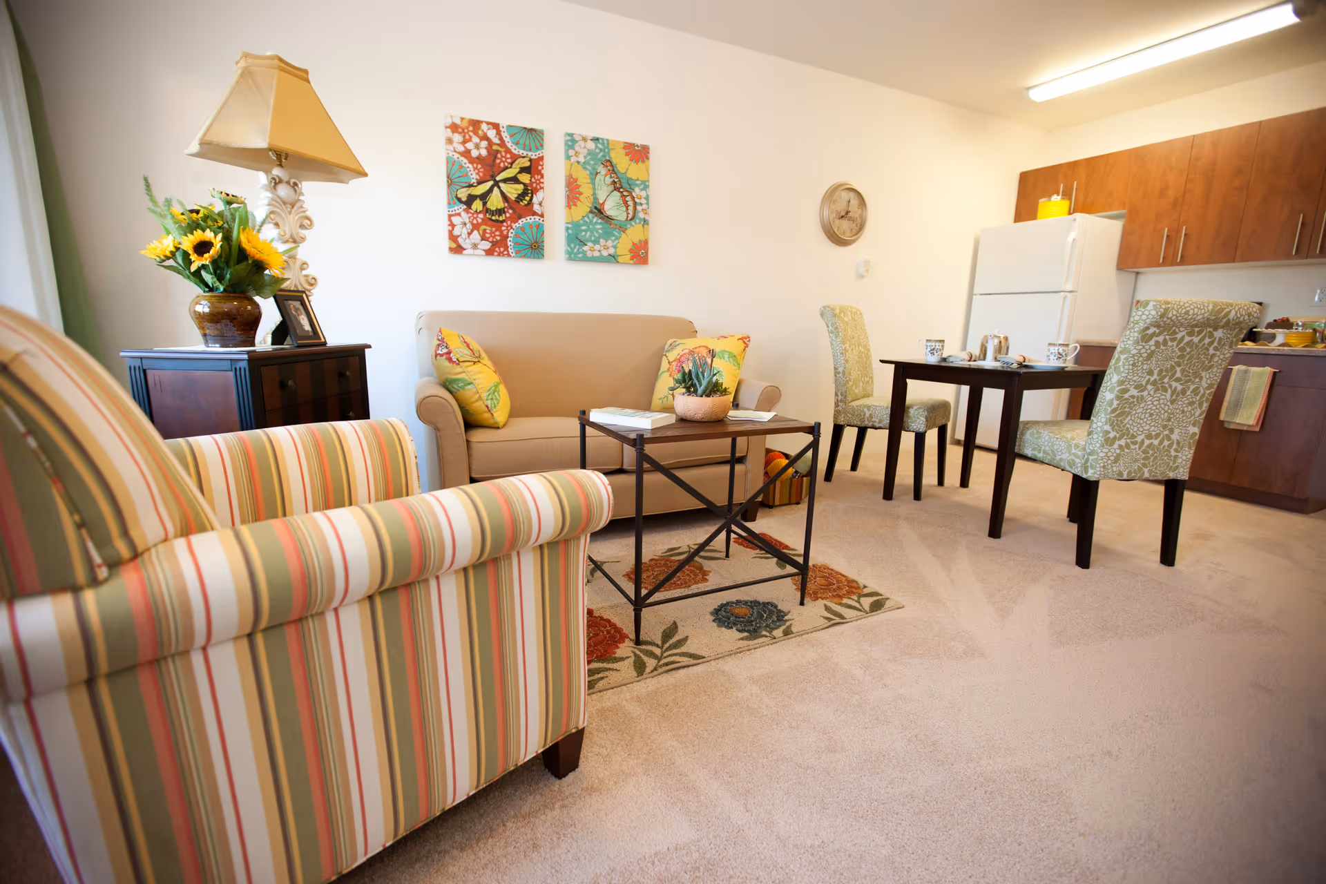 A cozy living area in a retirement facility featuring a striped armchair, a beige loveseat with butterfly-themed pillows, a small coffee table on a floral rug, a side table with a lamp and a vase of sunflowers, and a dining table with two green patterned chairs. In the background, there is a kitchen area with wooden cabinets and a white refrigerator.