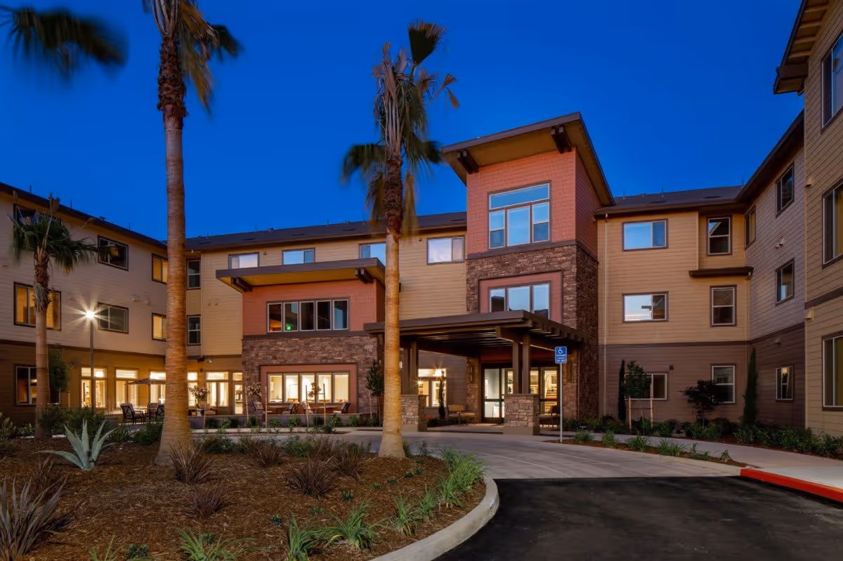 Exterior view of a three-story senior living facility building at dusk with illuminated windows, a covered entrance, palm trees, and landscaped garden beds in front.