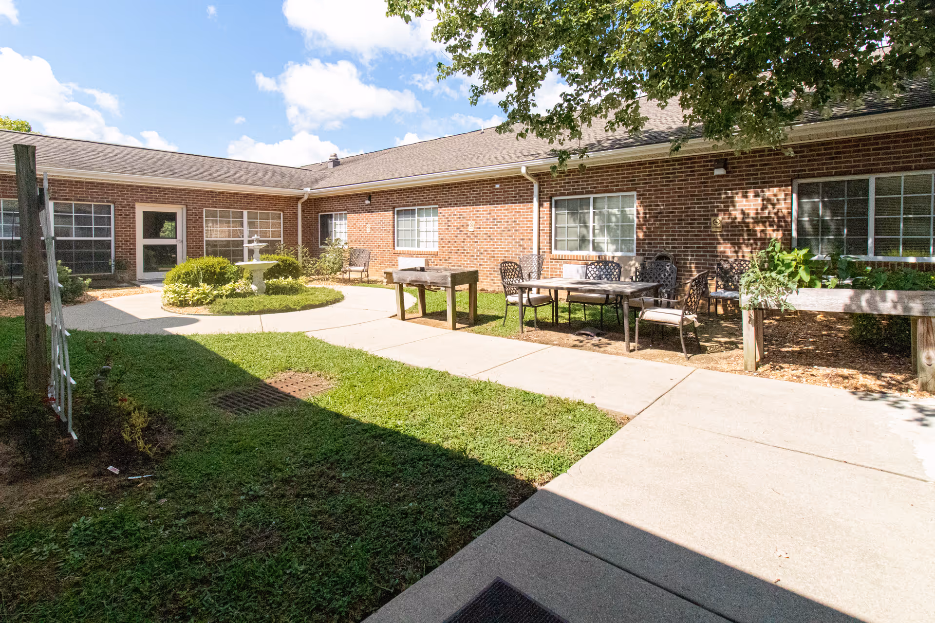 Sunlit brick courtyard with patio tables and chairs, a central fountain and planted greenery.