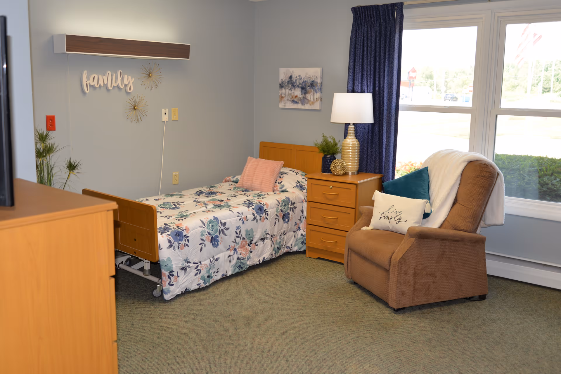 A furnished senior bedroom with a single floral bed, wooden nightstand and lamp, and a brown recliner by a large window.