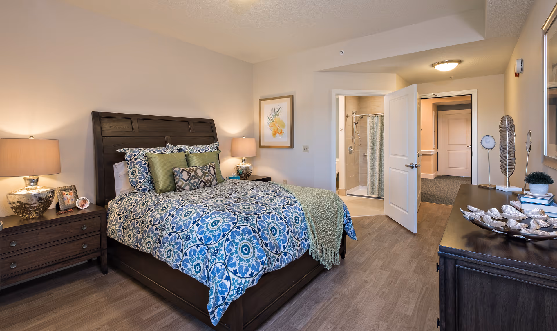 A well-lit bedroom featuring a dark wooden bed frame with a blue and green patterned comforter and multiple pillows. There are two bedside tables with lamps on each side of the bed. A framed floral artwork hangs on the wall above one bedside table. The room has light-colored walls and wood flooring. An open door reveals a bathroom with a shower and a curtain. A dark wooden dresser with decorative items is visible on the right side of the room.