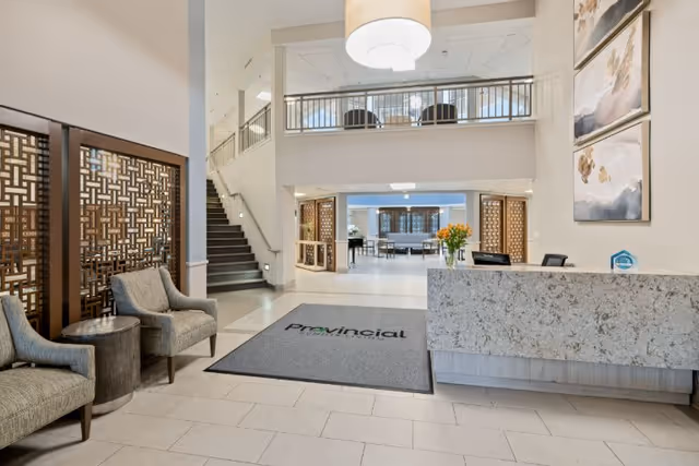 Spacious and well-lit reception area of a senior living facility with a modern stone front desk, two upholstered chairs with a small round table, decorative wooden partitions, a staircase leading to an upper floor, and a large hanging light fixture. The floor mat near the desk reads 'Provincial'.