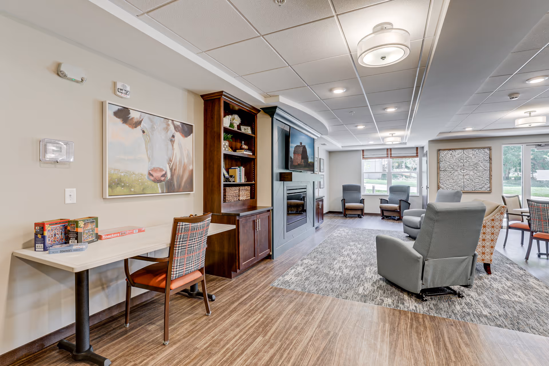 A bright and spacious common area in a senior living facility featuring a seating area with armchairs and a sofa arranged around a large fireplace with a TV above it. There is a table with board games and a chair on the left side, a large window letting in natural light, and decorative artwork on the walls.