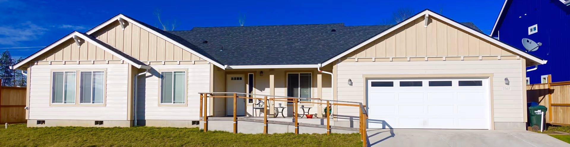 Single-story beige senior care facility building with a dark roof, a front porch with a wooden railing, a wheelchair ramp, and a two-car garage. The building is surrounded by a grassy lawn and a wooden fence under a clear blue sky.
