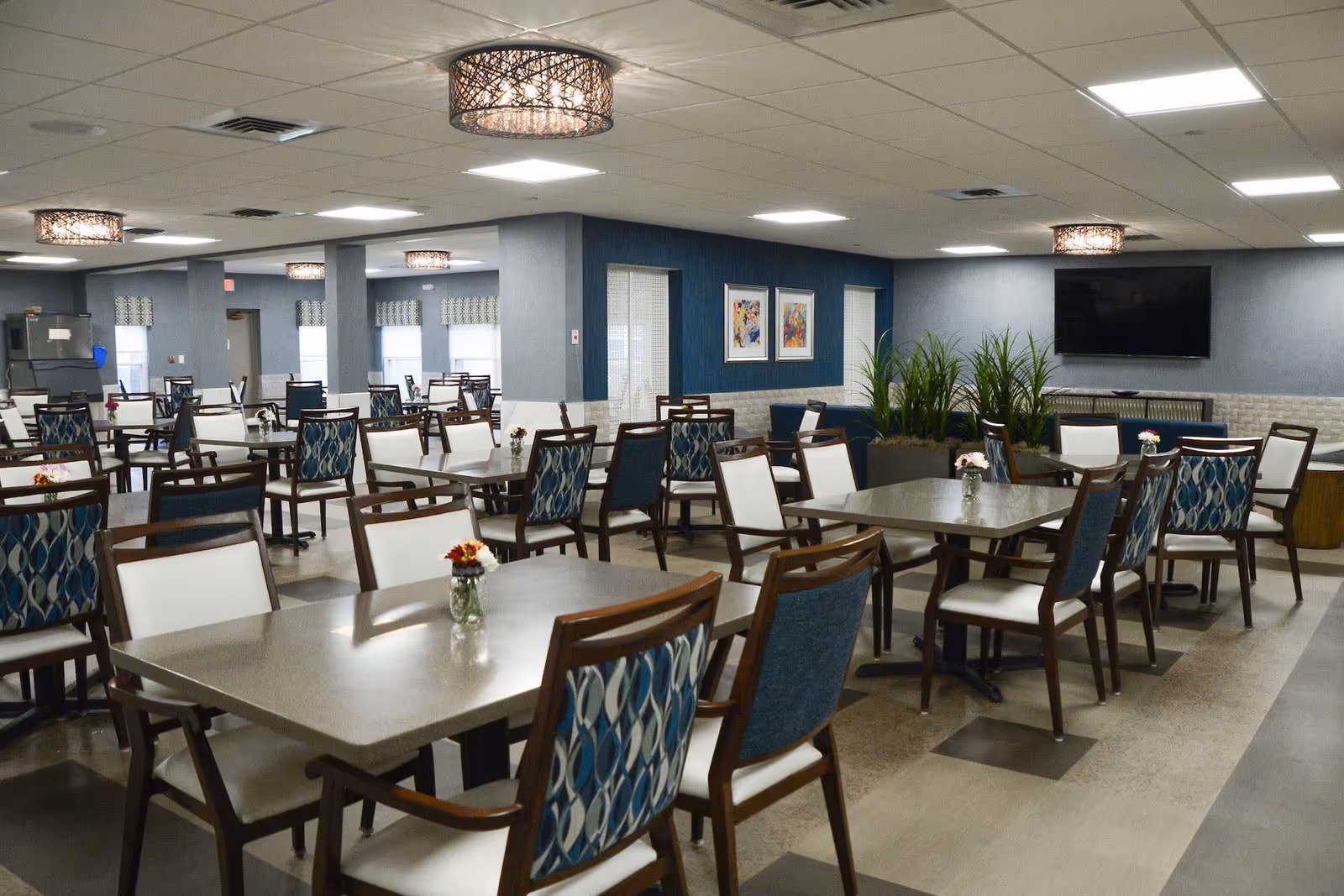 A spacious dining room with multiple tables and chairs arranged neatly. Each table has a small vase with flowers. The room features modern lighting fixtures on the ceiling, blue and white walls, framed artwork, and a large flat-screen TV mounted on one wall. There are also some green plants placed near the walls.