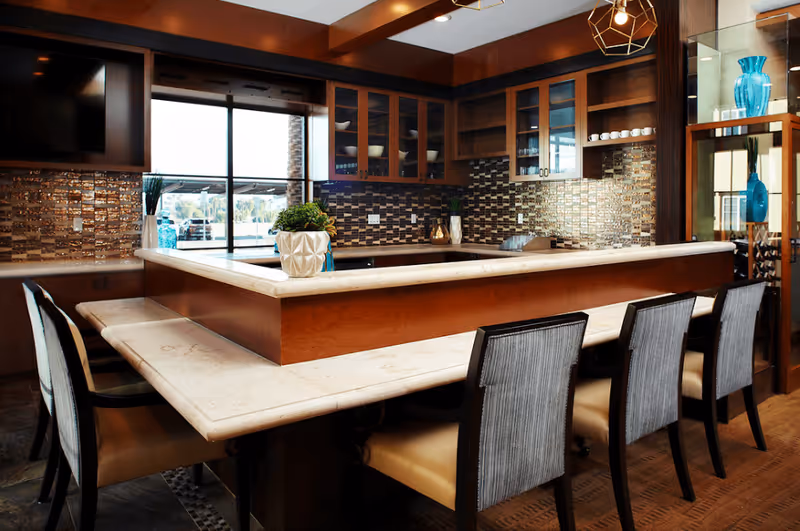 Modern kitchen area with a large marble countertop island surrounded by six chairs. The kitchen features wooden cabinets with glass doors, a tiled backsplash, and decorative vases. A large window provides natural light, and a flat-screen TV is mounted on the wall.