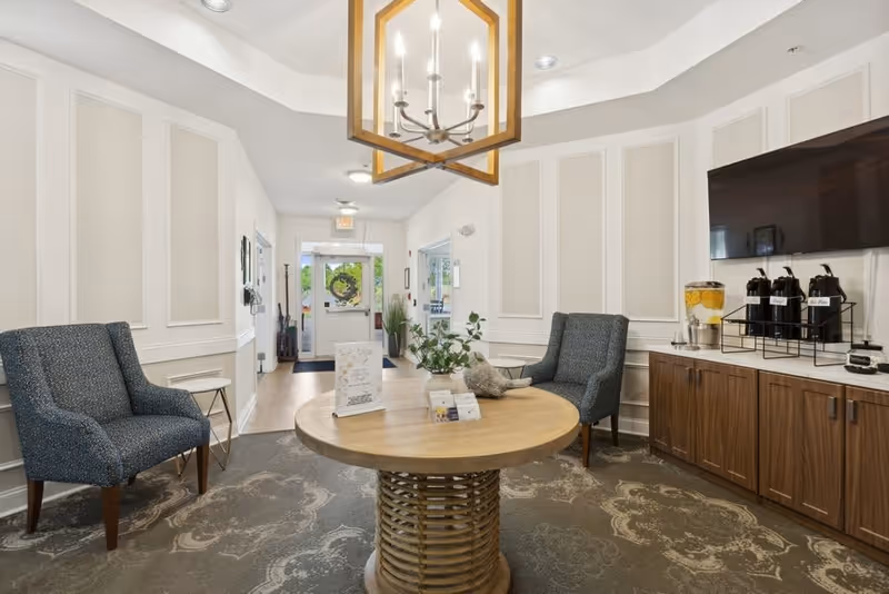 A bright and welcoming seating area in a senior living facility with two blue upholstered chairs, a round wooden table with informational brochures and a plant, a wooden cabinet with beverage dispensers, and a large flat-screen TV mounted on the wall. The space has white paneled walls, a patterned carpet, and a modern chandelier hanging from the ceiling. The entrance door with glass panels is visible in the background.