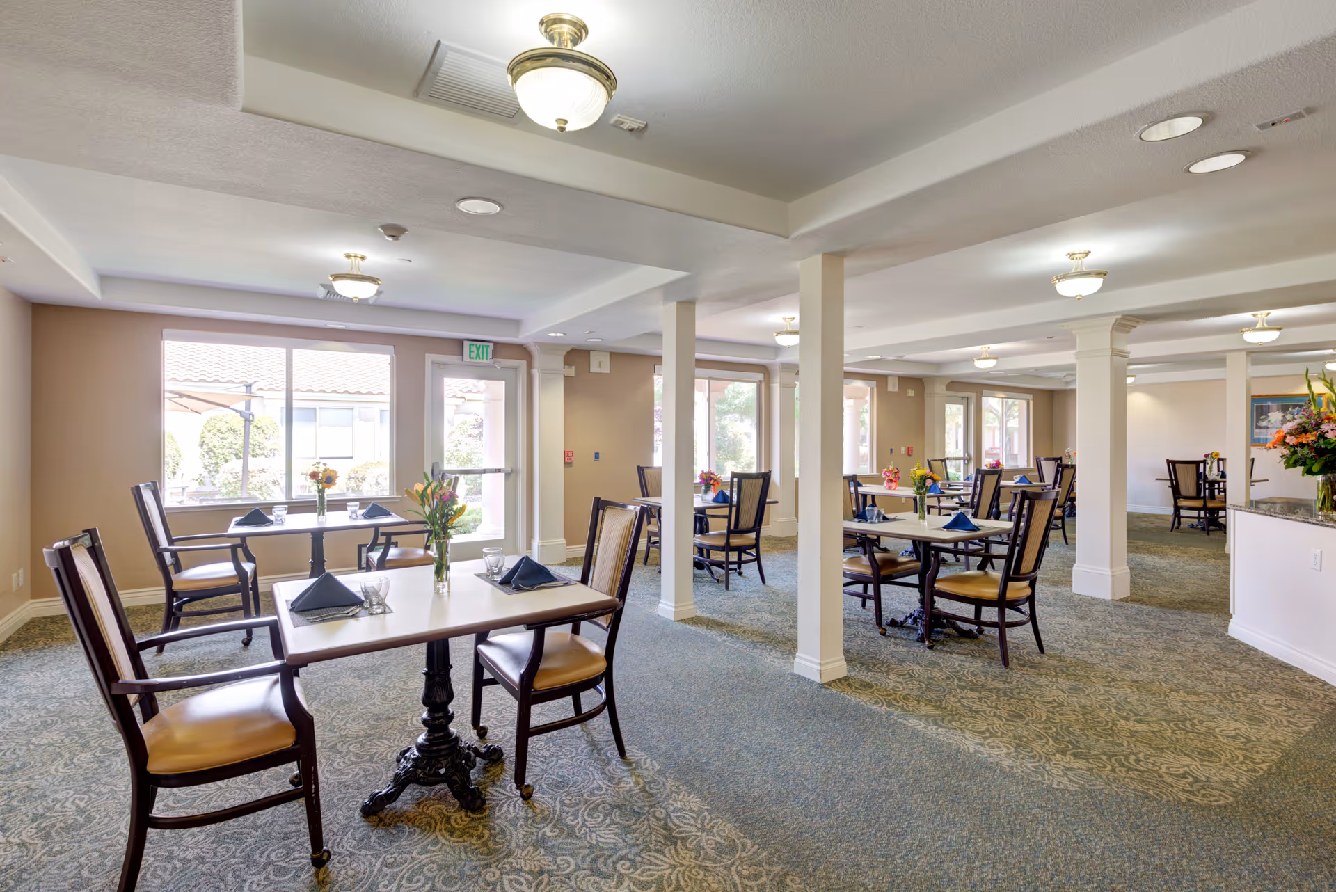 A bright dining room in a senior living facility with multiple tables and chairs arranged neatly. Each table has a small vase with flowers and folded napkins. Large windows allow natural light to fill the room, and there are several ceiling lights providing additional illumination. The room has a patterned carpet and beige walls.