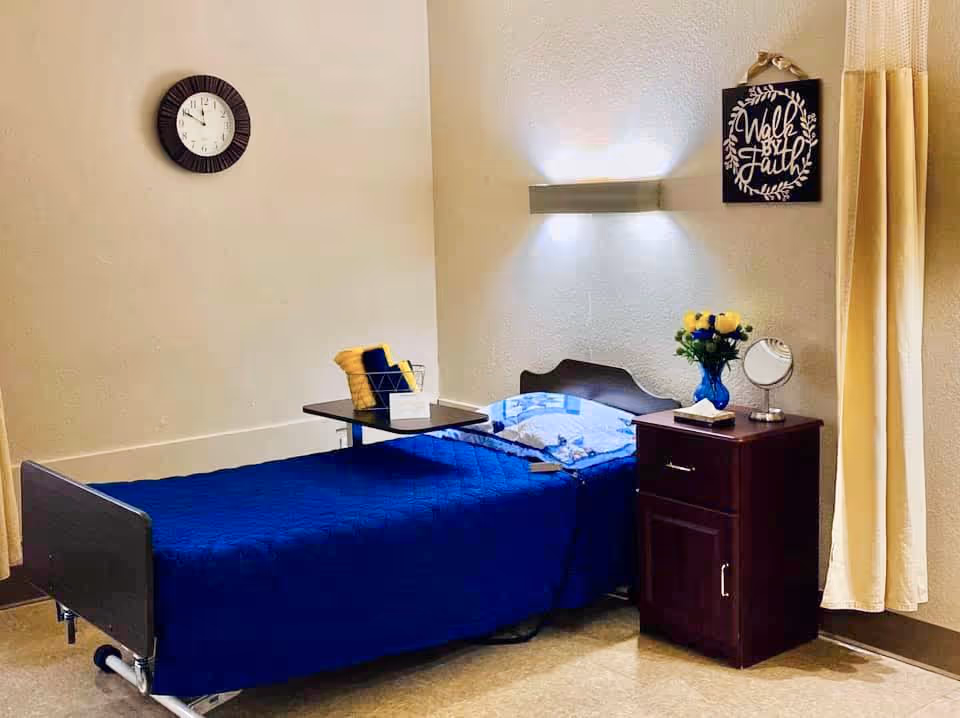 A small bedroom with a single bed covered in a blue quilt. There is a wooden nightstand next to the bed with a vase of yellow flowers, a small round mirror, and a book. Above the nightstand is a wall light and a decorative sign that reads 'Walk by Faith'. A clock is mounted on the wall above the bed, and a beige curtain hangs to the right side of the room.