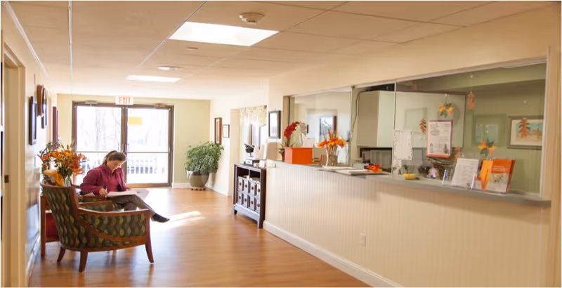 A bright hallway in a senior living facility with a woman sitting on a patterned armchair reading a book. The hallway features wooden flooring, a reception desk with a glass partition decorated with autumn-themed items, and large windows letting in natural light. There are plants and framed pictures along the walls.
