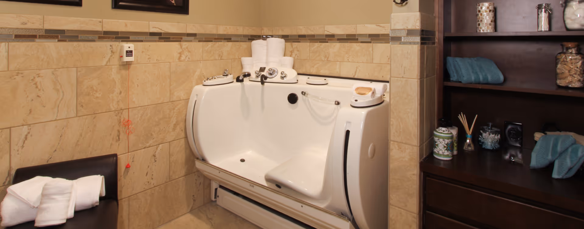 A beige tiled bathroom featuring a white walk-in bathtub with built-in controls and a seat. On the left, there is a dark chair with folded white towels on it. On the right, a dark wooden shelving unit holds various decorative items and folded blue towels. A red emergency pull cord hangs on the wall above the chair.