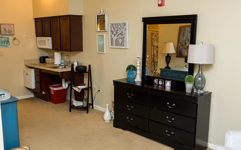 Studio-style interior showing a kitchenette with cabinets and microwave on the left and a black dresser with mirror, lamp, and decor on the right.