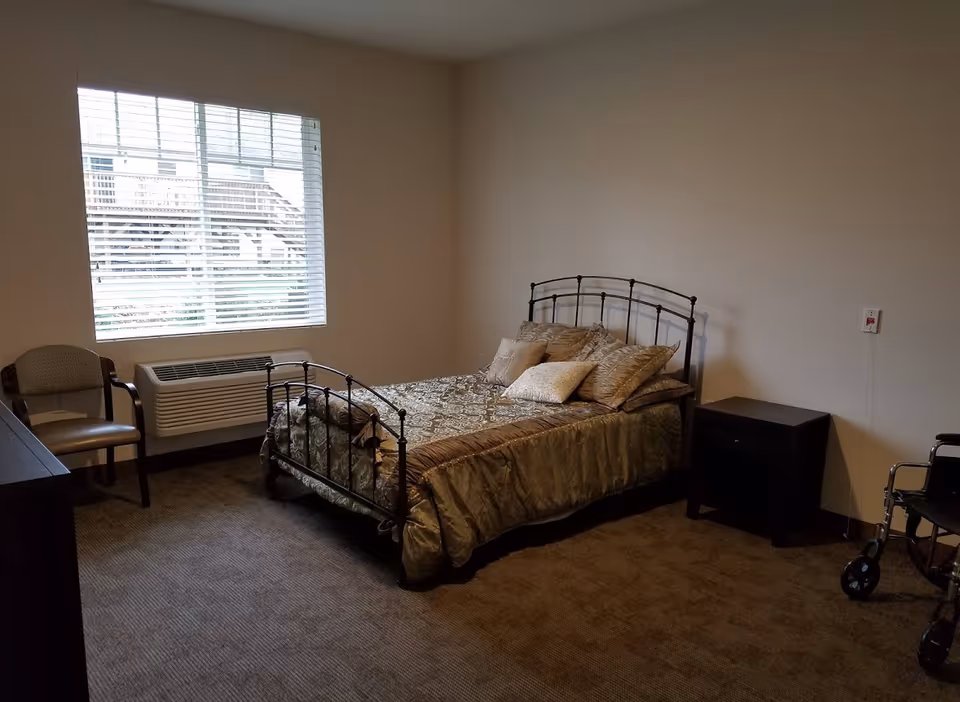 A modest senior living bedroom with a metal-framed bed, pillows, nightstand, chair, window with blinds, and a walker near the corner.