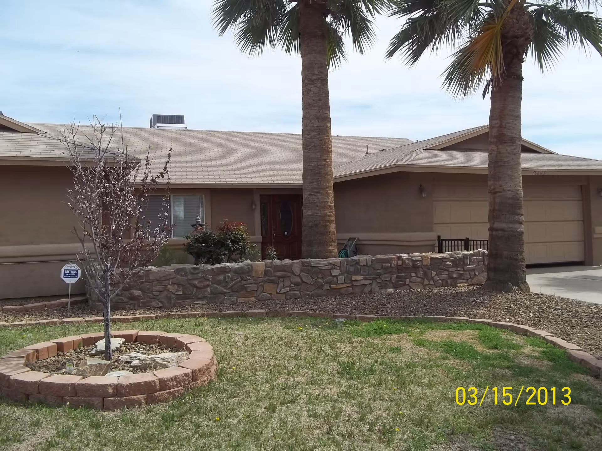 Front exterior view of a single-story house with a beige roof and walls, two tall palm trees in the front yard, a small circular brick planter with a young tree, a stone wall along the front of the house, and a driveway leading to a double garage door.