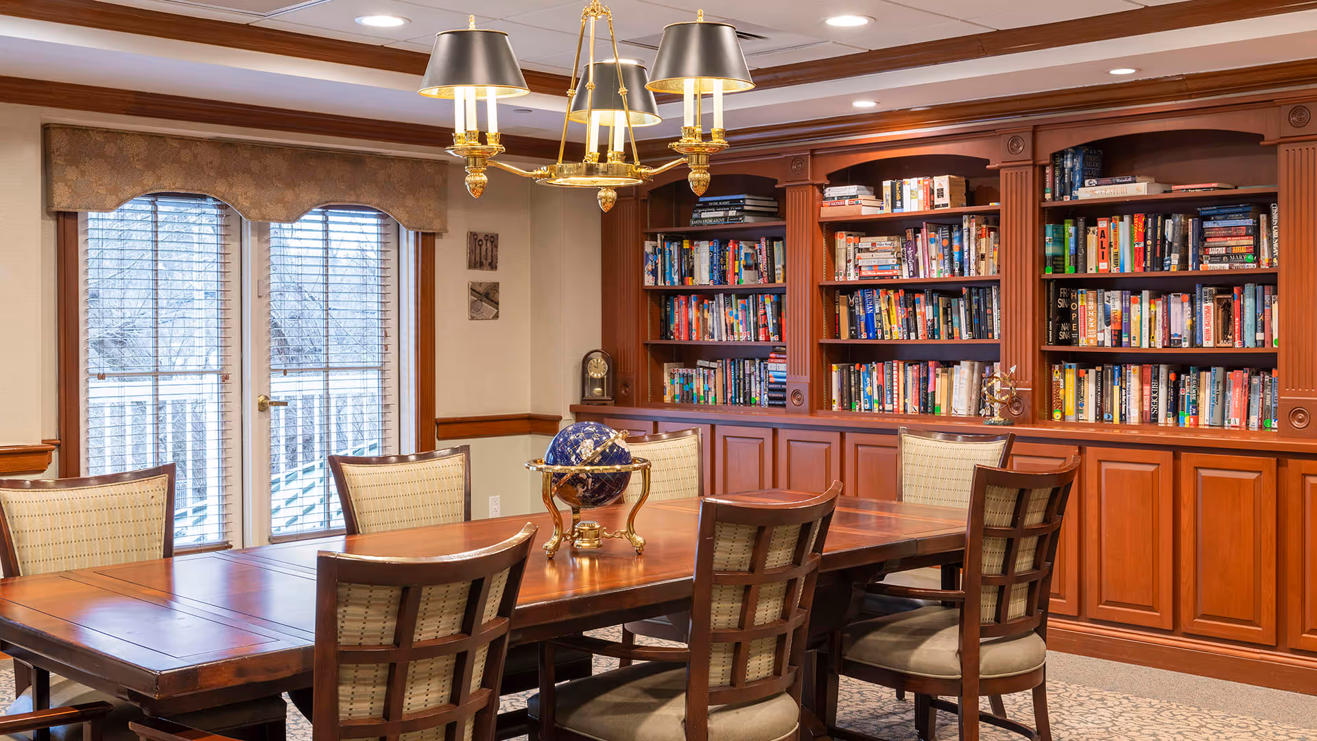 A cozy library room with wooden bookshelves filled with books along the back wall. In front of the shelves is a long wooden table surrounded by six cushioned chairs. A decorative globe sits on the table. Large windows with blinds and valances allow natural light into the room. A brass chandelier with black lampshades hangs above the table.