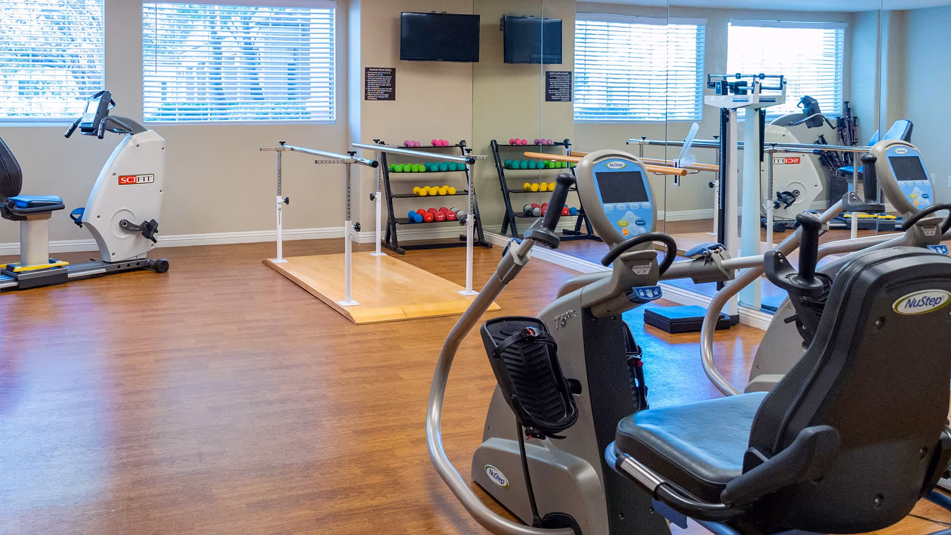 A fitness room with exercise equipment including NuStep recumbent cross trainers, a stationary bike, a rack of colorful dumbbells, a wooden platform with parallel bars, and large windows with blinds letting in natural light.