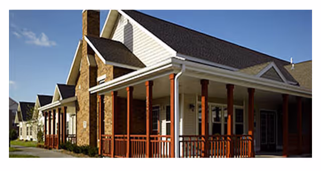 Front exterior of a senior living building with a covered porch, red wooden columns, brick chimney and pitched roof.