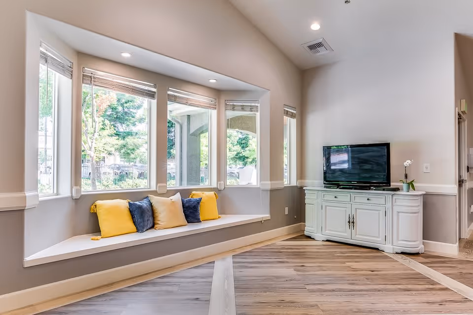 A bright and airy living room area with a large bay window featuring a built-in bench seat adorned with yellow and blue cushions. Across from the window is a white cabinet with a flat-screen TV on top and a small potted plant. The room has light-colored walls, recessed lighting, and wood flooring.