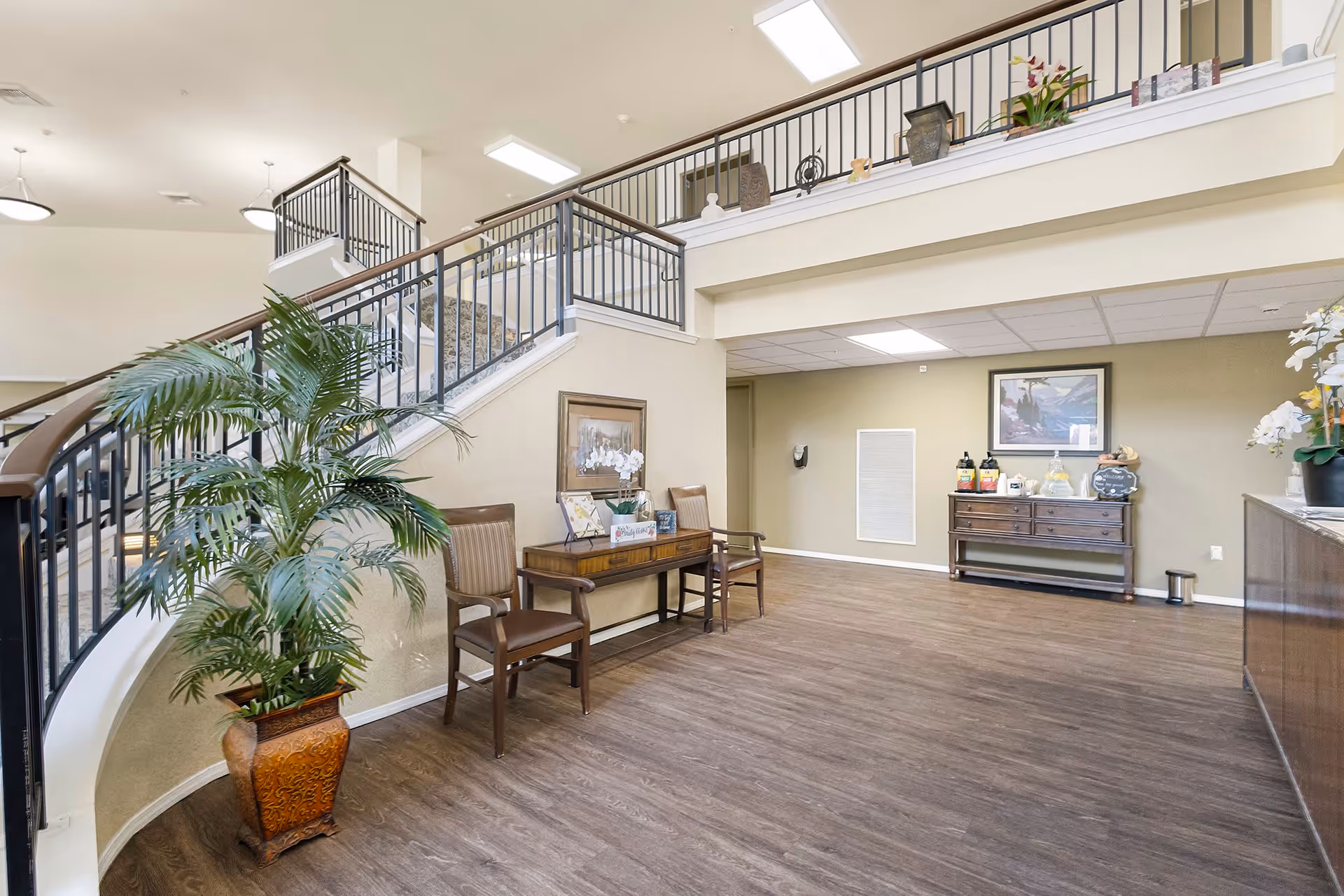 Interior view of a senior living facility lobby area with a staircase featuring black railings and wooden handrails. There is a large potted plant near the staircase, two wooden chairs with a small table between them, and a sideboard with refreshments and a framed picture on the wall behind it. The floor is wood, and the ceiling has recessed lighting.