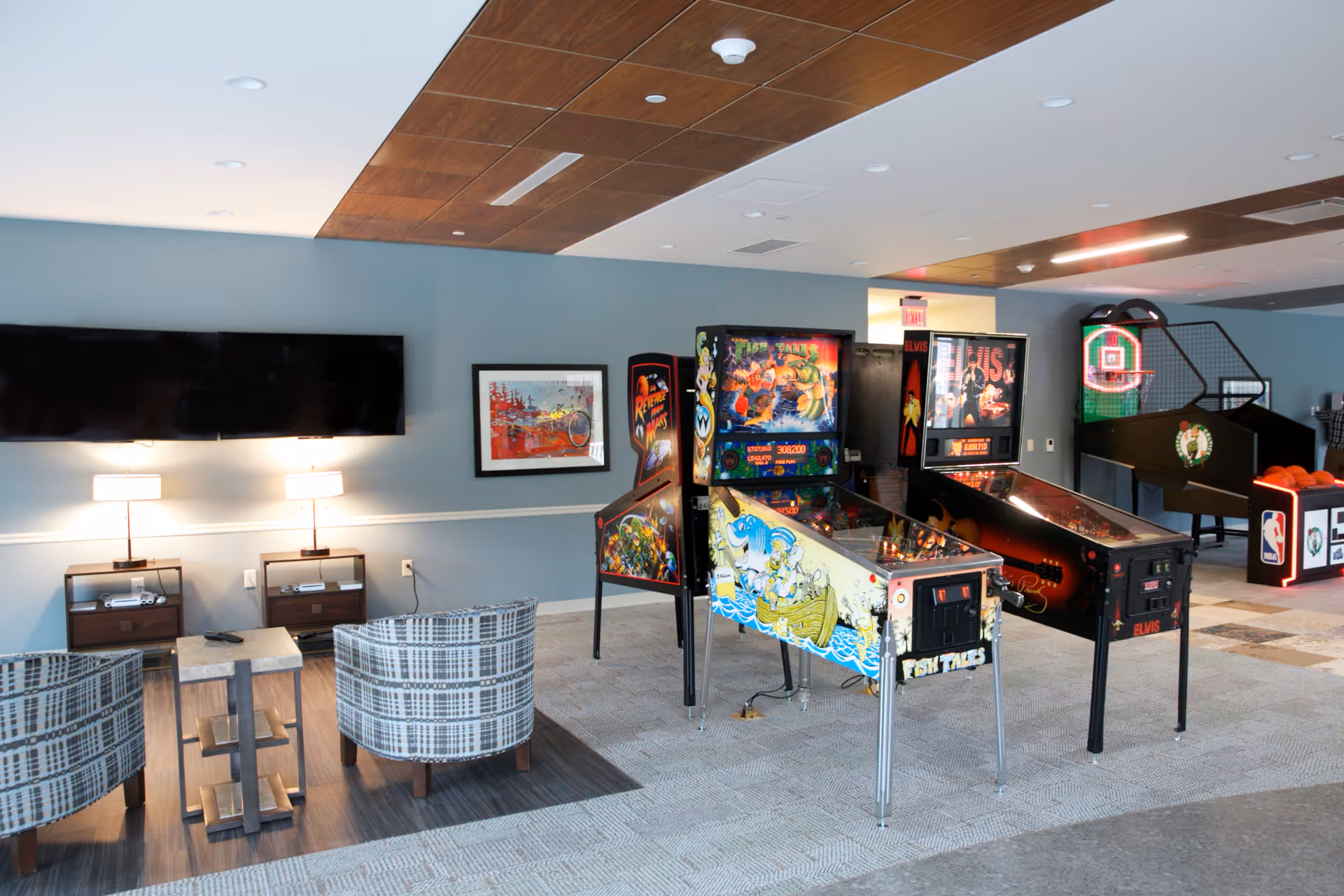 A recreational room with two pinball machines and a basketball arcade game. There are two patterned armchairs facing a wall-mounted flat screen TV with two small tables and lamps underneath. The room has a modern design with a wooden ceiling panel and light blue walls.