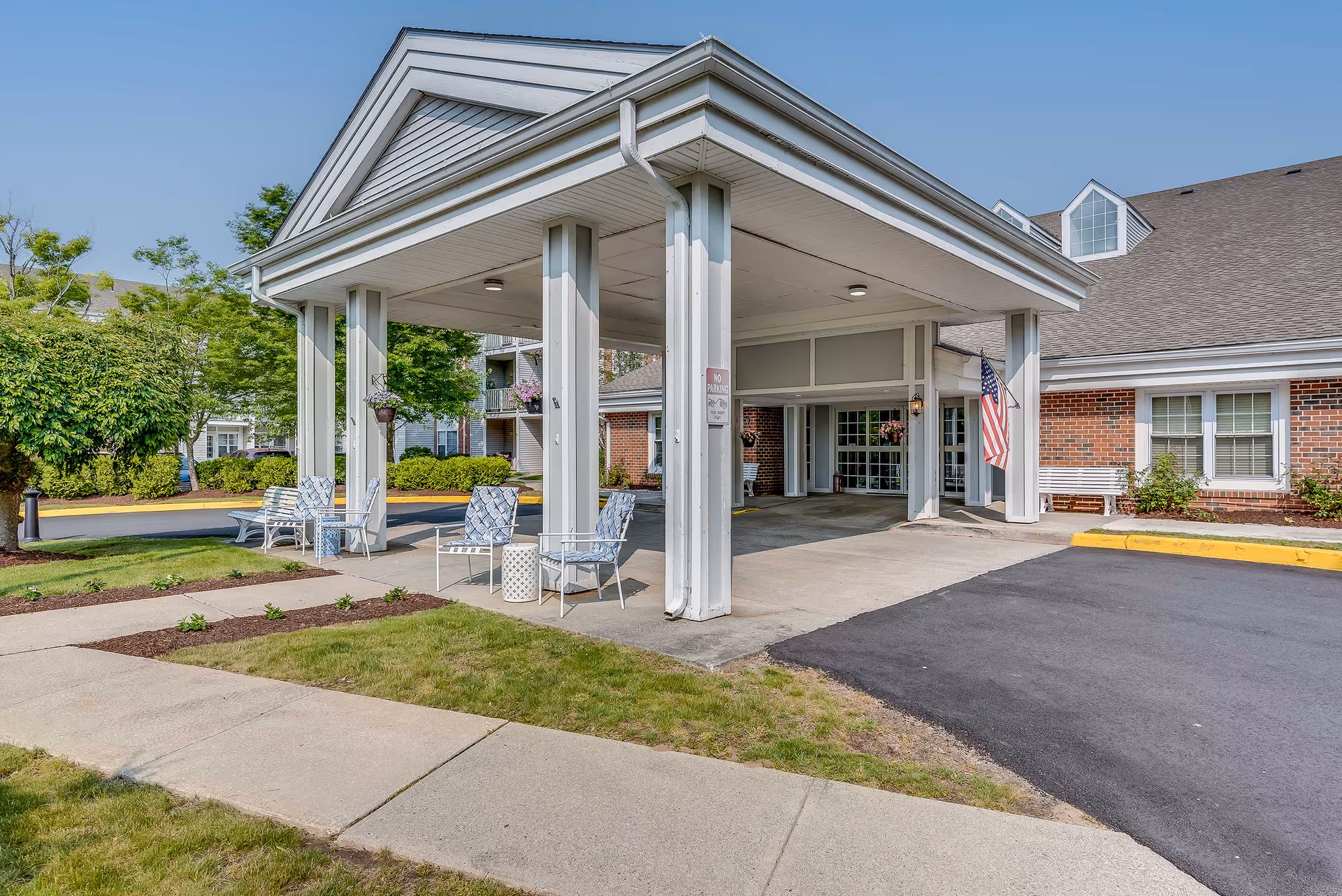 Covered porte-cochere entrance of a senior living facility with outdoor chairs, hanging plants, and an American flag.