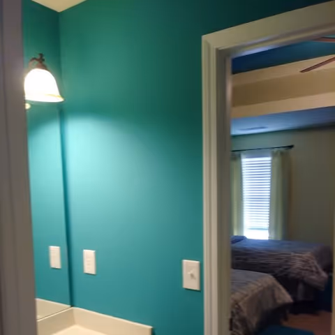 View of a teal-colored wall with a lit wall-mounted lamp and two light switches, next to a doorway leading into a bedroom with two beds covered in gray bedding and a window with closed blinds and curtains.