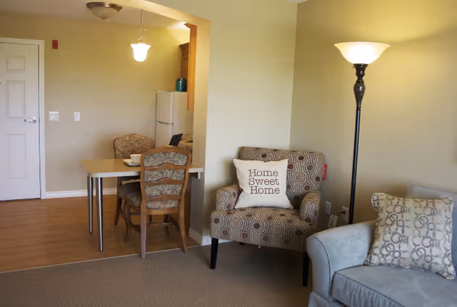 A cozy living area with a patterned armchair featuring a 'Home Sweet Home' pillow, a floor lamp, and a light blue sofa with a patterned cushion. In the background, there is a small dining table with two chairs and a kitchen area with a refrigerator.