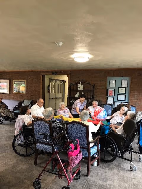 A group of elderly people sitting in a circle in a common room, some in wheelchairs, engaging in a group activity with a colorful beach ball. A caregiver is interacting with them. The room has carpeted floors, framed pictures on the walls, and a ceiling light fixture.