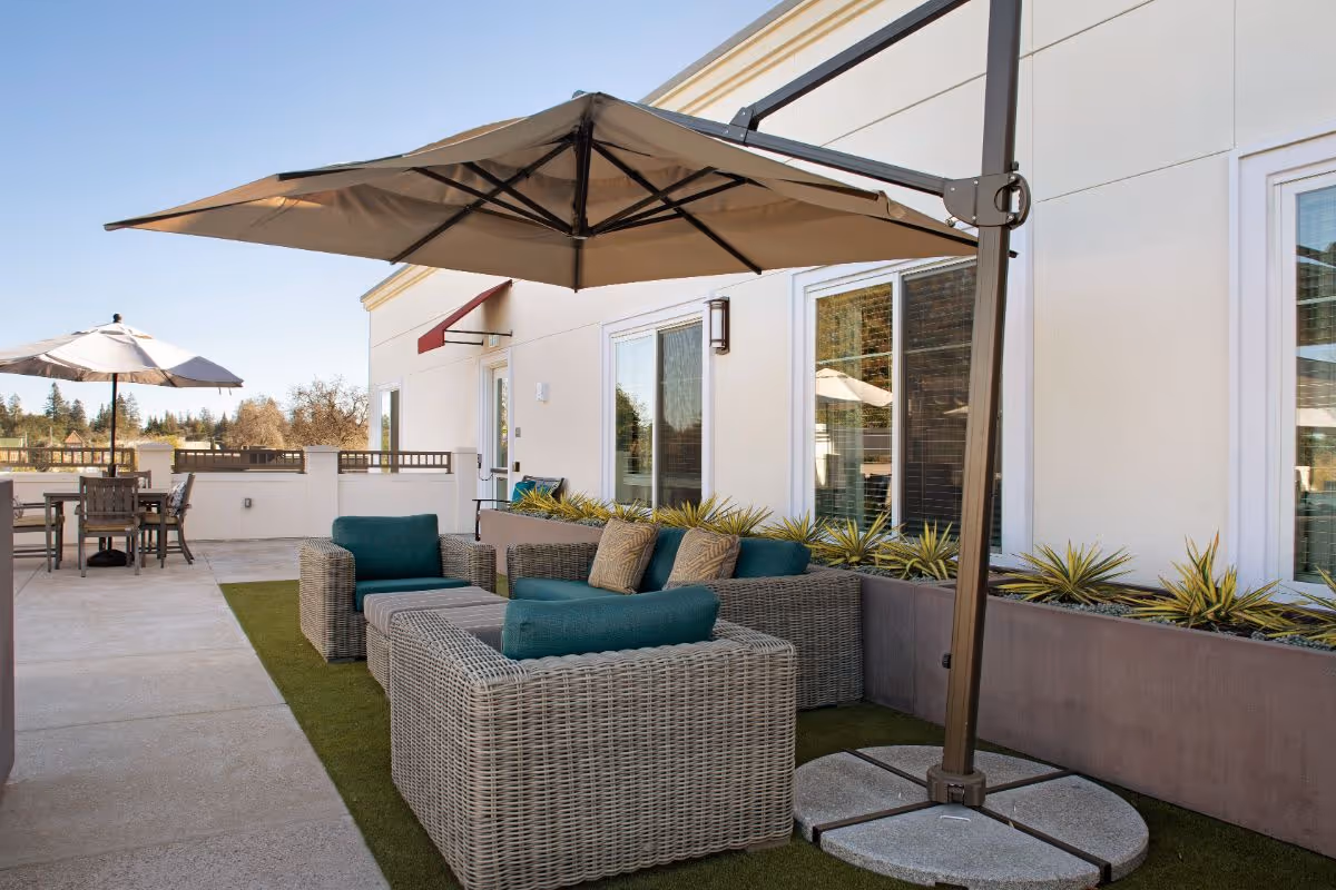 Outdoor patio area at Oakmont of Redwood City with wicker seating furniture featuring teal cushions, a large beige umbrella providing shade, and a table with chairs and another umbrella in the background. The patio is adjacent to a building with windows and planter boxes containing small plants.