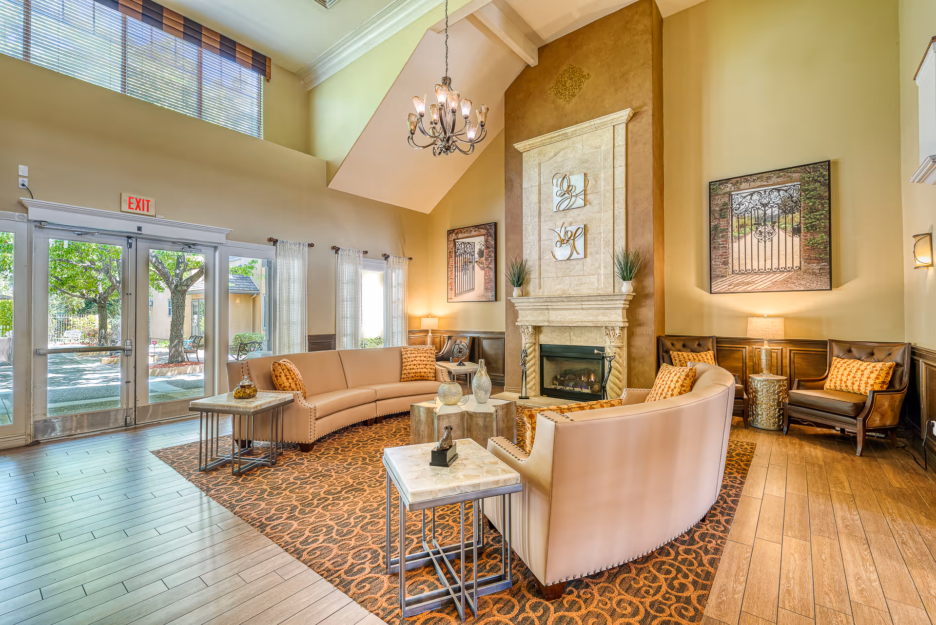 Bright, elegant senior living lounge with curved sofas arranged around a fireplace, chandelier, and large windows/doors to the outside.