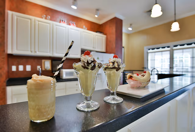 A kitchen counter with a milkshake in a mason jar with a striped straw, two ice cream sundaes topped with whipped cream and cherries in glass dishes, and a banana split dessert on a white plate. The background shows white cabinets, a microwave, and a window with blinds.