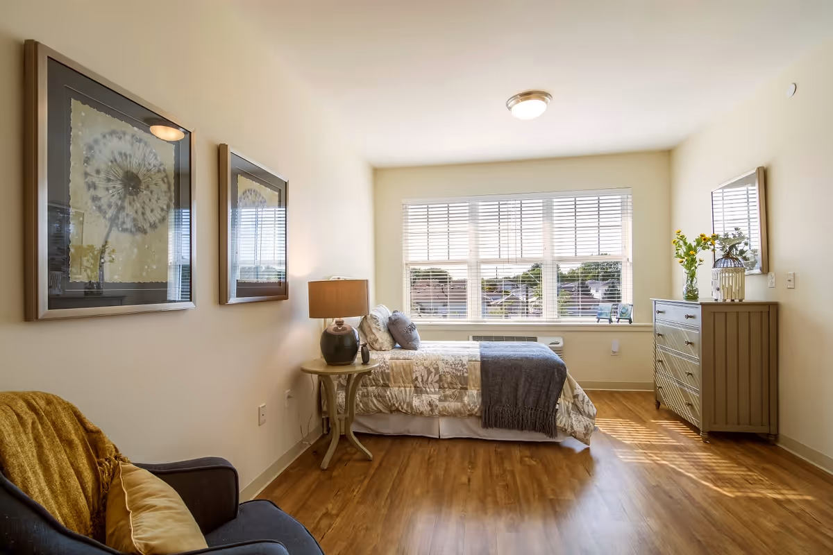 A bright and cozy bedroom with a single bed covered in a patterned quilt and a gray throw blanket. There is a wooden nightstand with a lamp next to the bed, and a wooden dresser with a mirror and decorative items on top. Two framed dandelion prints hang on the wall above a dark armchair with a yellow throw blanket and pillow. Large windows with white blinds let in natural light, illuminating the room's wooden floor.
