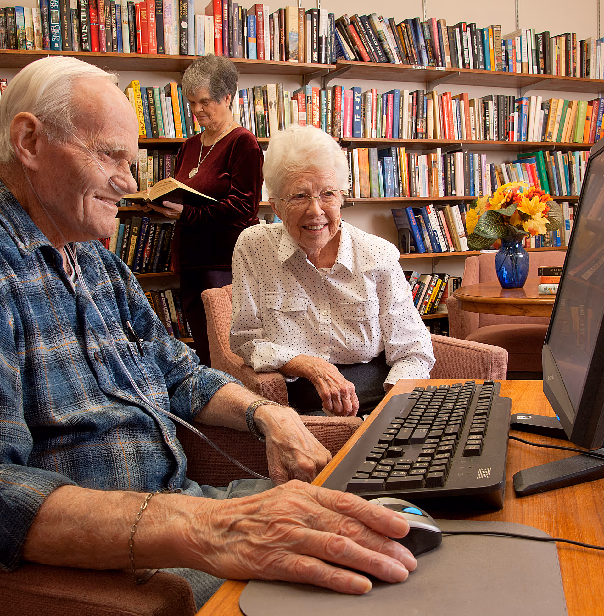 Three elderly people in a library/common room with bookshelves: a man using a desktop computer (with a nasal cannula), a smiling woman beside him, and another woman reading a book in the background.