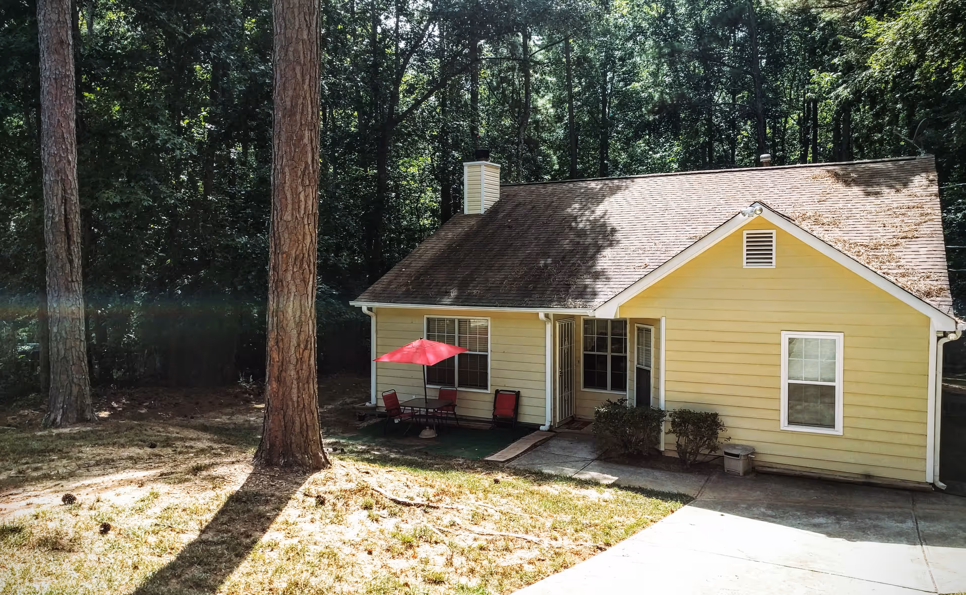 A small yellow house with a sloped roof surrounded by tall trees. There is a concrete driveway leading up to the house, and a small patio area with a red umbrella and outdoor chairs. The house is set against a backdrop of dense forest.